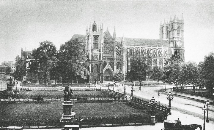 A black and white photograph postcard of Parliament Square. In the centre and just in the background is Westminster Abbey, with the west towers to the right of the picture, and the north rose window above the entrance in the centre. In front of the Abbey in the foreground is Parliament saqure, separated by a wlkway through the middle towards the abbey's entrance. To the left standing in a grassy recangular area is a statue of Robert Peel, behind this area is a similar grassy area whihc holds two more statues. 