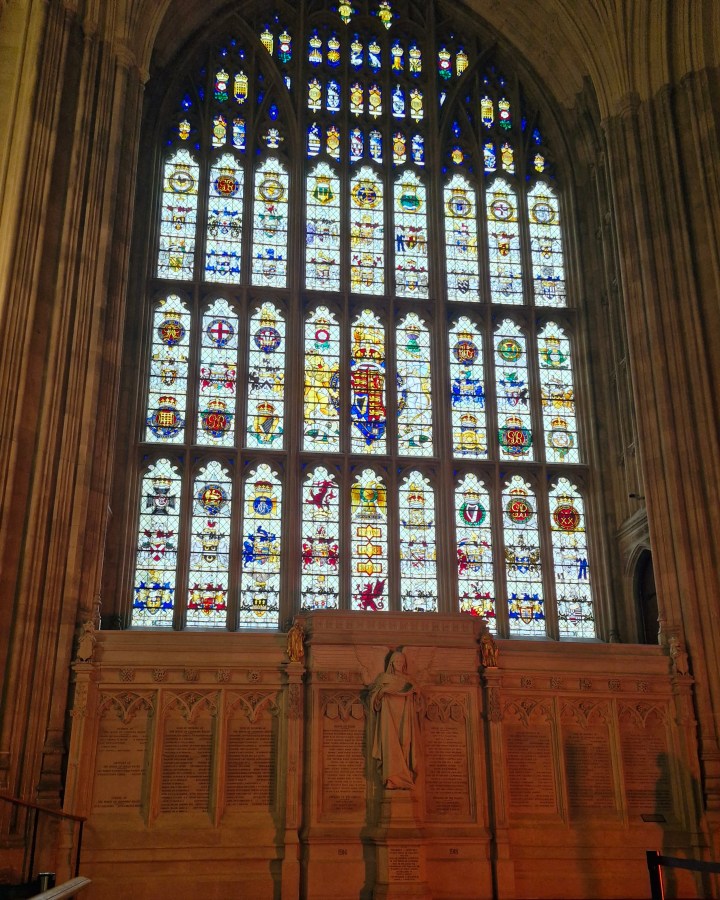 A picture of the Recording Angel memorial in Westminster Hall. With an angel statue in the middle, either side engraved in stone tablets in a large memorial wall are the names of MPs, peers, officers and their sons who lost their lives in the First World War. Above the memorial is a very tall stained glass window adorned with crests.