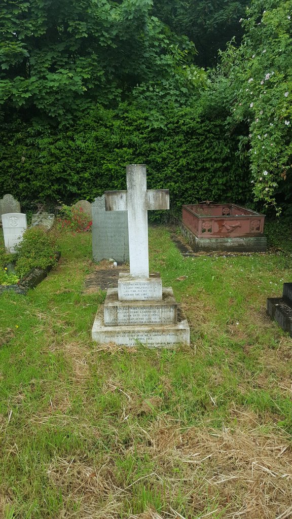 A picture of William Glynne Charles Gladstone's grave. In the middle of the picture stands the grave with a white cross on top, with a three tiered plinth with text on. It is surrounded by green grass and behind the grave is a darker green hedge.