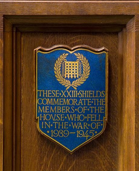 A blue painted shield against a wooden panelled background. It has gold lettering which reads THESE XXIII SHIELDS COMMEMORATE THE MEMBERS OF THE HOUSE WHO FELL IN THE WAR OF 1939-1945.