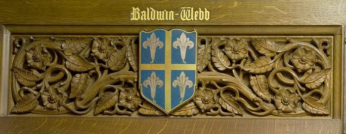 A carved wooden panel with a design of leaves and flowers. In the centre is a blue shield with a coat of arms. It is in four quarters separated by a gold cross and each quarter contains a gold fleur de lis emblem. The lettering above the shield reads Baldwin-Webb.