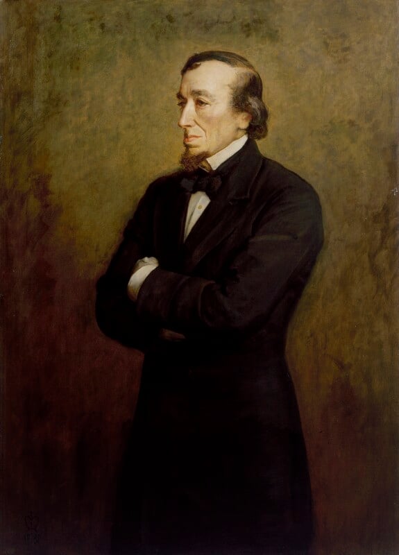 A knee-length portrait of Benjamin Disraeli. Standing half side on, behind a background which fades from darker brown in the bottom left hand corner to a paler cream in the top right, he is wearing a black suit with a white shirt and black bowtie. He has a protruding goatee, with combed receding greying hair.  