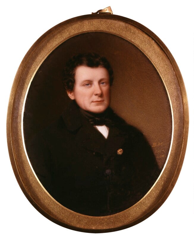 A half-length portrait of Daniel O'Connell in an oval bronze frame, In front of a brown background, he is wearing a black suit jacket, with a small gold button next to the lapel of the jacket, a white shirt and brown necktie. He is clean shaven with short brown hair. 