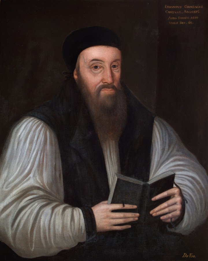 Half portrait of a man sat with a black book open in his hands. He has a long nose and a long brown beard. He is wearing traditional clerical costume of a white robe with long black waistcoat-style layer over his shoulders and a black hat on his head.