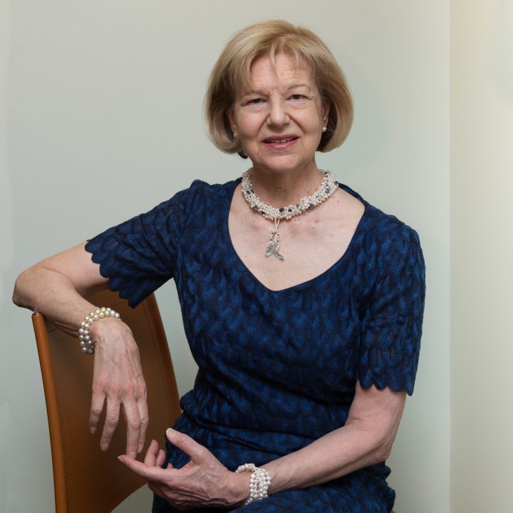 Photograph portrait of a Emma Nicholson. Nicholson is sat side-on on a wooden chair, her right arm resting on the back of the chair, while her left arm rests in her lap. She has blonde shoulder length hair, and is wearing a dark blue dress and a decorative pearl necklace, bracelet and earrings. 