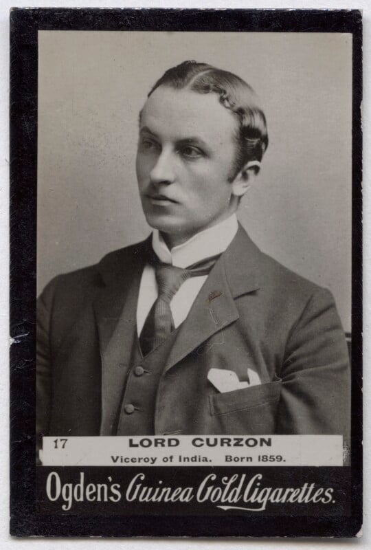 A half-length black and white photographic portrait of George Curzon. Looking to the left of the image. he is weating a dark suit coat with a pale handkerchief poking out of his jacket pocket, a dark waistcoat, dark tie with a white collared shirt. He is clean shaven with short combed side parted hair.