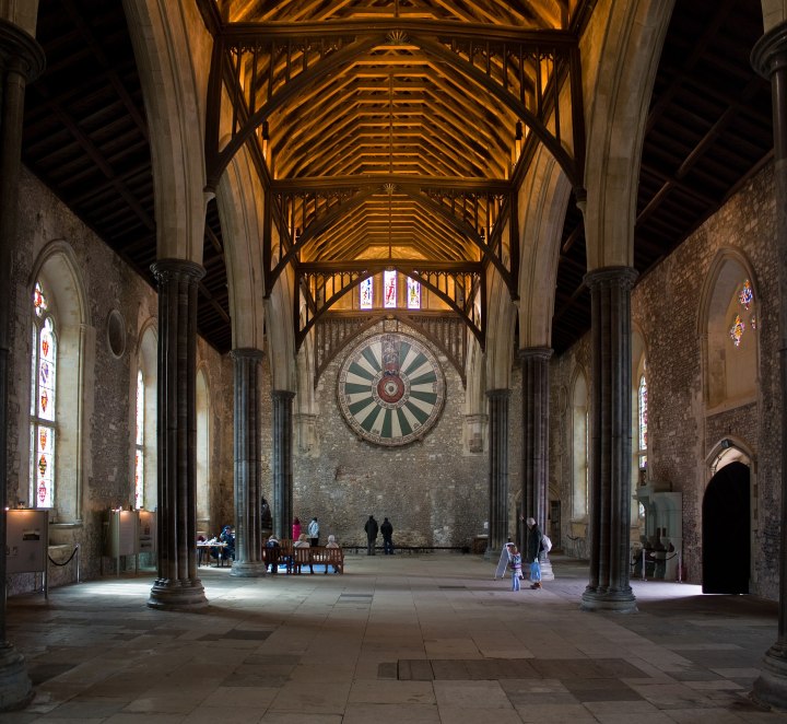 A large medieval hall, with arched windows in the walls, and a tall wooden roof supported by two rows of stone columns.