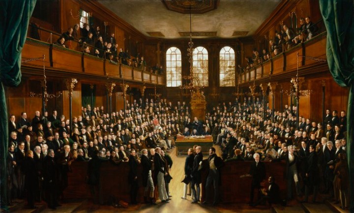 Painting of the interior of the House of Commons chamber. There are three arched windows at the back of the room. It is a large open space with benches for seating and there are also seating galleries on the upper level on either side. The room is crowded with men, mostly sitting on the benches, but some standing. A chandelier hangs from the ceiling in the centre and beyond this there is a decorated chair for the Speaker.