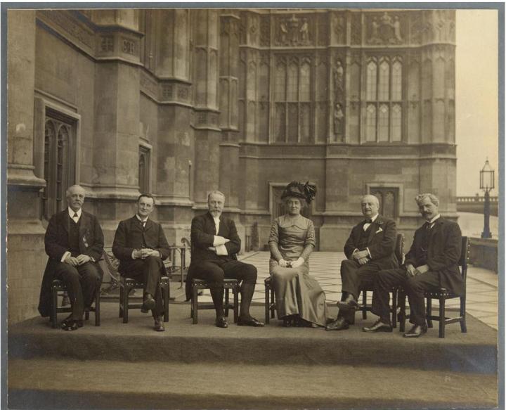 Six people (one woman and five men) sitting on chairs on a terrace outside the UK Parliament, with Parliament and the River Thames in the background.