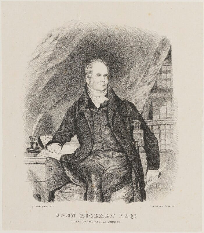 A man in nineteenth-century full suit, sitting with his hand resting on a document on a writing table, with a quill in his hand.  In the background to his right is a bookcase, partly covered by a drawn curtain. The caption at the bottom reads John Rickman esq., clerk of the house of commons.