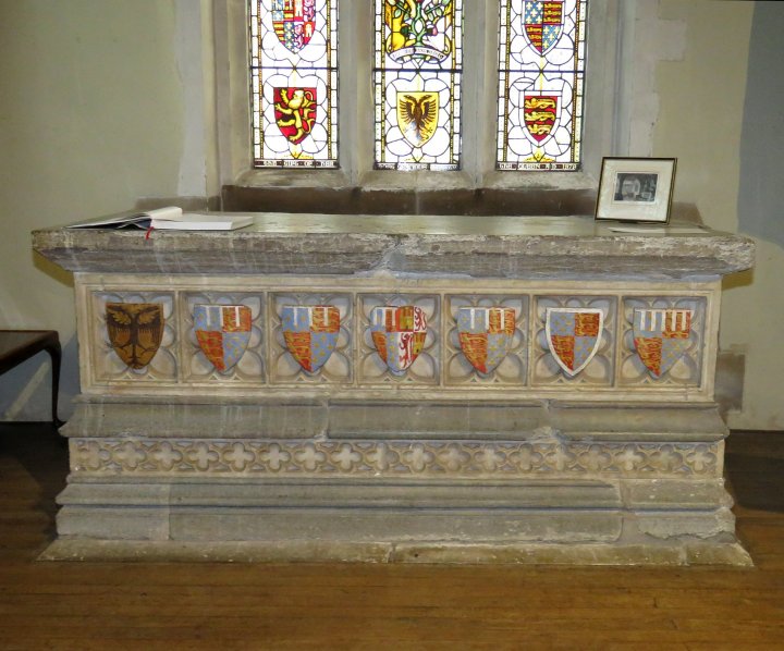 A coloured picture of the tomb of Edmund duke of York and Isabella of Castile. Inside on a wooden floor is the tomb decorated with quatrefoil horizontally across the tomb. Above this are seven coloured coats of arms in a line. Above and behind the tomb is a stained glass window also decorated with coats of arms.