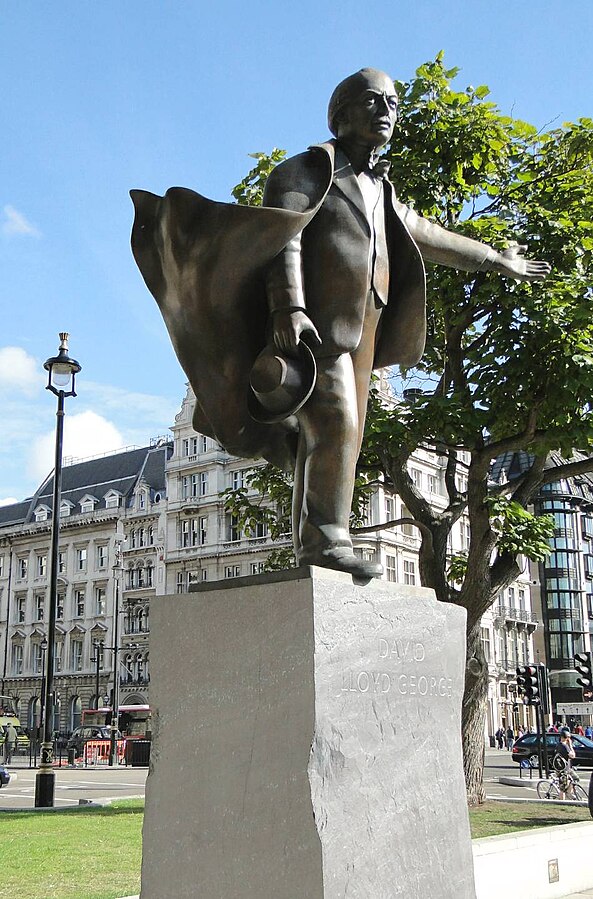 A coloured photograph of David Lloyd George's, statue in Parliament Square. On an imperfect cube stone plinth with his name carved into the stone stands Lloyd George, gesturing with his left hand off to the left, and holding his hat in his right down by his side. He is wearing a suit with a bowtie, with his jacket billowing behind him in the wind. He is clean shaven with short swept back hair. 