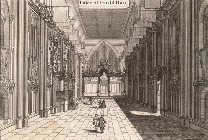 Engraving of the view inside London's Guildhall. Interior of a hall, with flags along the ceiling, and five figures walking towards the end of the room. 