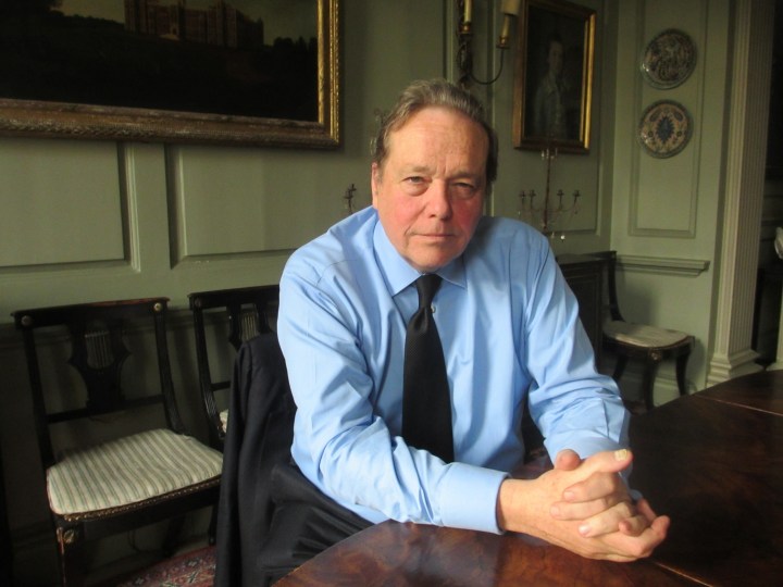 A photograph is portrait of Lord Salisbury. Sitting at a wooden table with his hands clasped together placed on the table, he is wearing black suit trousers, a pale blue shirt and a black tie. His suit jacket is hung on the back of his chair. He is clean shaven with brown combed hair. The wall behind Salisbury is a light green, with wooden white a green striped upholstered chairs lining the wall. There are two pictures hung on the wall both with golden frames. 
