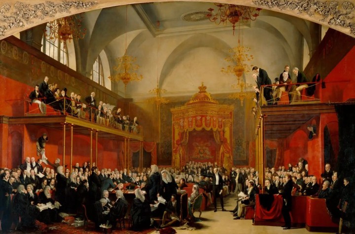 A painting of the House of Lords chamber during the trial of Queen Caroline. The room has a high vaulted ceiling with six golden chandeliers hanging. The two side walls are decorated red and on the back wall sits the Throne of Great Britain, decorated ornately in red and gold. The room is full of peers, mostly sitting but some standing and addressing the front. There are two balconies on either side wall also full of attending peers. Those at the front are sitting at a table in wigs sorting through stacks of paper. Just to the right of them in a small green chair sits Queen Caroline.