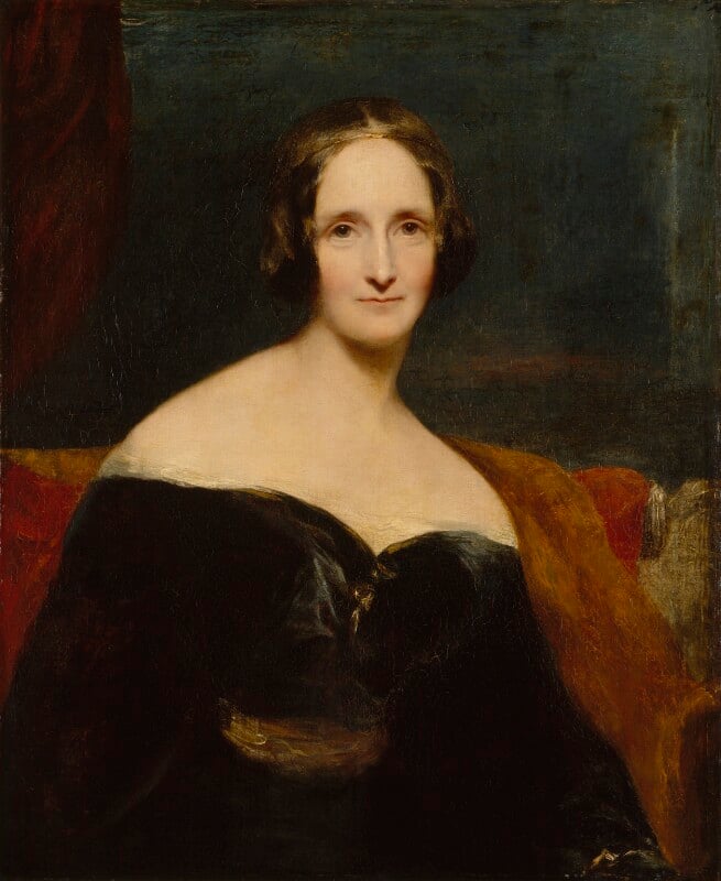 An oil painting of author Mary Shelley from above the waist. Shelley is seated and is wearing a black long-sleeved dress with a sweetheart neckline which sits just off her shoulders. Shelley is seated on an orange-red seat in front of a dark background. She looks directly at the artist with her hair framing her face above her chin.