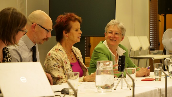 Three women and a man sit in a row at a table. They are listening to the red haired woman in the middle. The woman at the end is wearing a green jacket and smiling at the woman talking. 