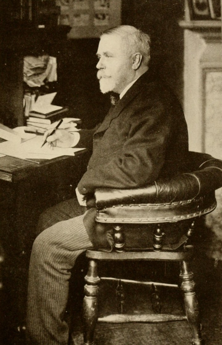 A black and white photograph of a man. He has white hair and a white beard and moustache. He is sitting at a desk in a leather chair with a low curved back. He has a pen in his hand and appears to be writing.