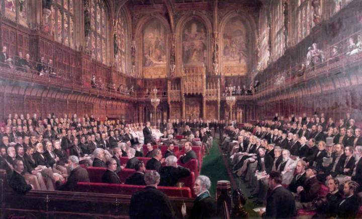 A painting of the House of Lords in the late 1800s. From the perspective at the back of the House looking to the Sovereign's chair at the far end, the House is full of sitting peers, mostly all dressed in black suits, with a small section to the far left dressed in white. They are filling the benches to the left and right and also the red chairs facing the front. There are two viewing galleries over each side of the chamber, each with a few people watching, with some women overlooking the house to the right. 