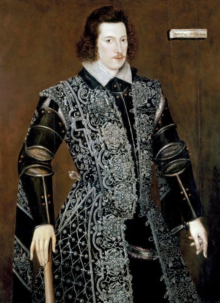 three quarter portrait of a man stood leaning against a staff. He has very pale skin and curly brown chin length hair. He has a whispy moustache. The man wears black breeches and top, with black armour style sleeves, and a tabard covered in pearls and silver embroidery.