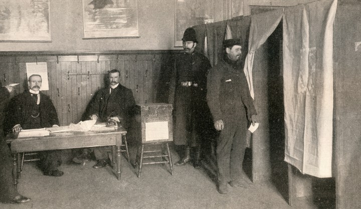 A black and white photograph depicting the voting procedure after the secret ballot in Glasgow. Two men are sitting at a table with papers in front of them. To the right of them is a large ballot box on a stool, with an officer standing next to it. To the right of the picture, a man is waiting to enter one of the four booths covered by a curtain for privacy, with a slip in hand to cast his vote secretly. 