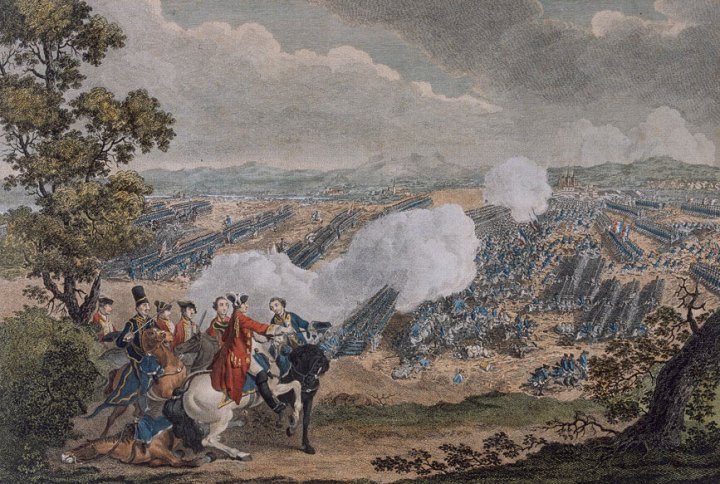 A coloured line engraving of the Battle of Minden.In the foreground on the left next to a shrub and a tall thin tree, there are military men on horseback, some in blue uniform and some in red. In the background in a wide open yellowing field depicts a large force of soldiers in formation in blue uniform. In the middle of the background a large portion of the army looks decimated with solders out of formation either engaging or on the floor, with plumes of white smoke around them.  