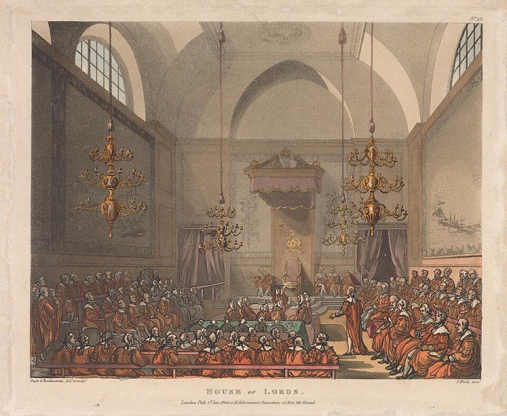 A painting of the House of Lords from the early 1800s. From the perspective from the back of the room looking towards the empty Sovereigns chair at the far end, which is a small chair with a light red canopy over the top. Those in arttendance fill the House, all wearing their red robes of the British peerage. There are four golden chandeliers hanging low over the attendees from the high vaulted ceiling of the room. One man standing from the right bench is speaking to the room. 