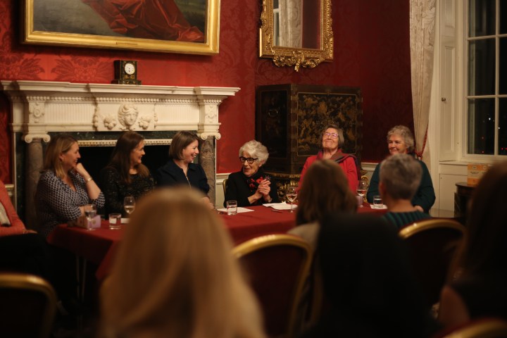 Six women are sat in a row at a table covered in a red table cloth. 