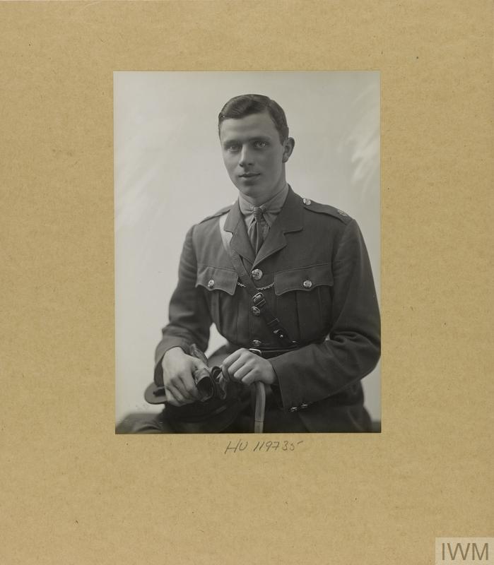 Half length black and white photograph of a young man dressed in military uniform. He appears to be seated and is holding the curved handle of a walking stick in one hand, and a pair of gloves and a cap in the other.