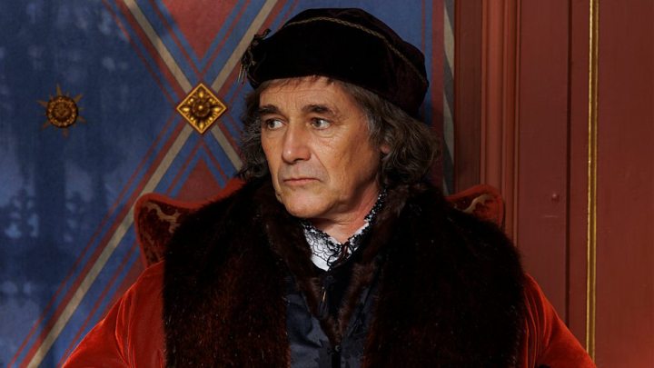 Photo still of a man dressed as Thomas Cromwell. He is sat in front of a red and blue patterned wall. He is wearing a white shirt with black lace collar details, and a large coat with fur collar time. He has a flat black hat on his head. 