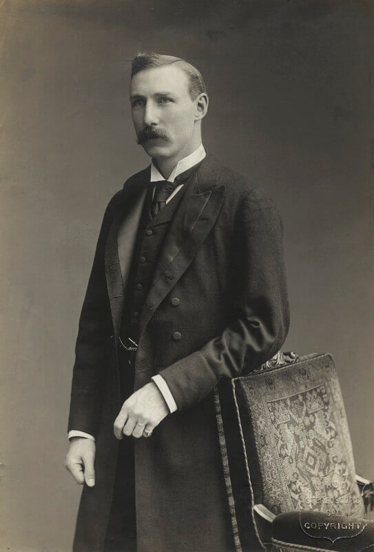 A three-quarter-length photographic black and white portrait of William Waldegrave Palmer. Standing in front of a blank background, with his left arm resting on a decorated cushioned armchair, he is wearing Victorian dress, with a dark long suit jacket open, a dark waistcoat with a dark tie and white collared shirt. He has short side parted combed hair and a thick moustache.  