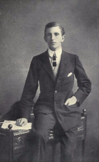 A black and white photograph of William Glynne Charles Gladstone. He is a young man wearing a full black suit with a white shirt and black tie. He is clean shaven with his hair combed to the left. He is leaning on a writing desk.