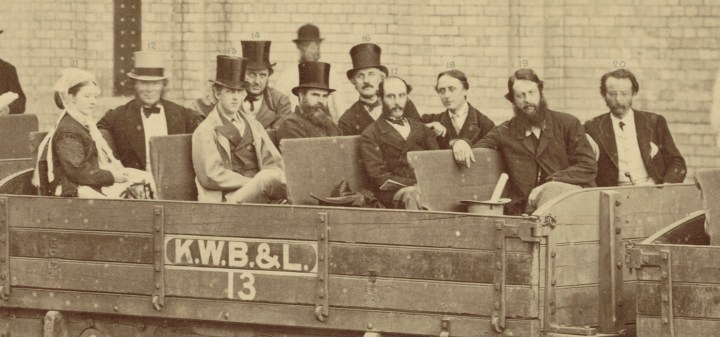 A yellowed black and white photograph of a group of dignitaries sat in an open wooden carriage on the metropolitan railway. The carriage reads on the front 'K.W.B.&L. 13'. There are four rows of dignitaries and 10 in total. There are nine men and one women, who sits at the far left of the picture. Lord Ronald Gower sits in the second row from the left, visually distinct from the rest of the people in the carriage wearing darker suits, as Gower is wearing a light overcoat. 