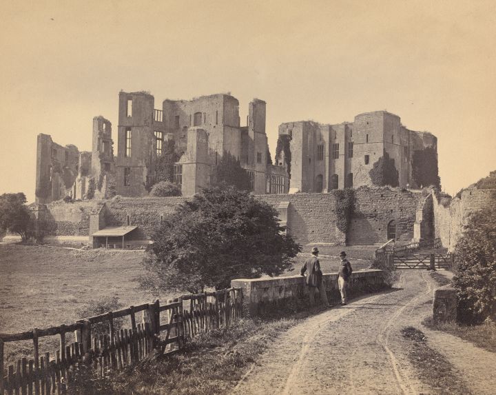 A black and white landscape photograph of Kenilworth Castle. In the foreground to the right is a path moving upwards towards the castle, with a wooden fence and then low wall separating the path from a field. Two men in bowler hats are standing by the wall talking. Above in the background stands the castle elevated from the field below with a high stone wall. The castle is delapidated with no roof and many parts of the castle walls having fallen down. But there are still a few windows in tact. 